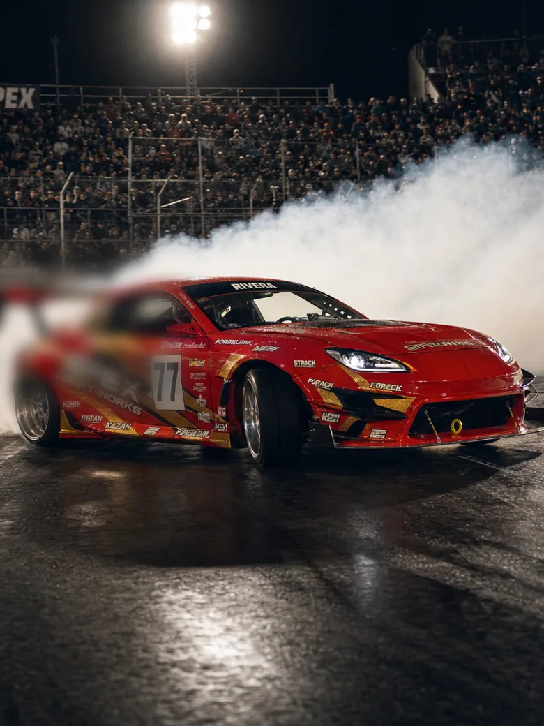A packed night drift event: light towers, grandstand crowd, a large LED queue display, a merchandise tent and cars mid-corner in tyre smoke